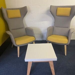 Two ergonomic chairs with high backs and yellow accents, positioned around a small white table with wooden legs