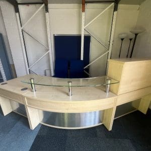 Modern office reception desk with a curved design, glass partition, and storage units, surrounded by chairs and lighting