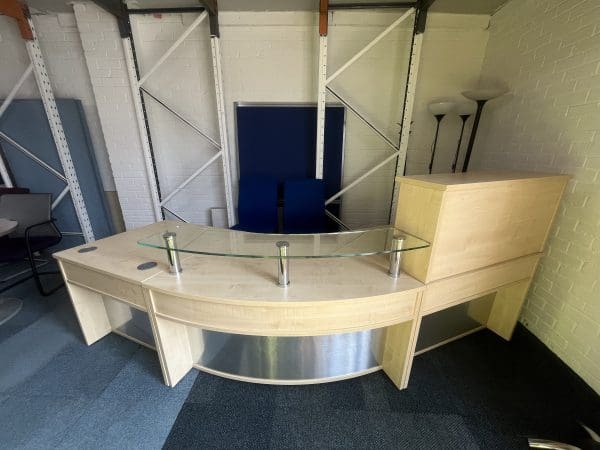 Modern office reception desk with a curved design, glass partition, and storage units, surrounded by chairs and lighting