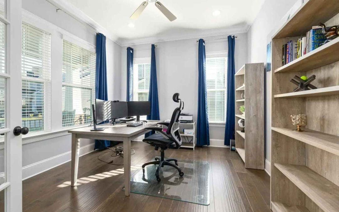 A bright modern home office with a desk, ergonomic chair, dual monitors, and wooden bookshelves filled with books and decor, featuring large windows with blue curtains.